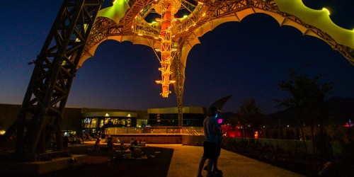 A vibrant structure illuminated at night, with people enjoying the space beneath it in a lively outdoor setting.