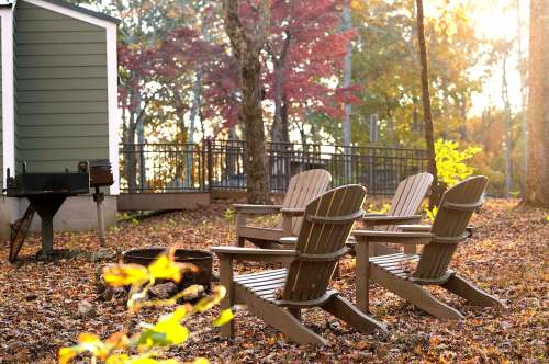 Four wooden Adirondack chairs sit in a forested area with autumn leaves, near a grill and a cabin in the background.