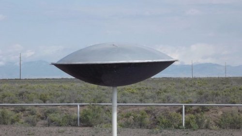 A metallic, saucer-shaped object on a pole in a desert landscape with mountains in the background.