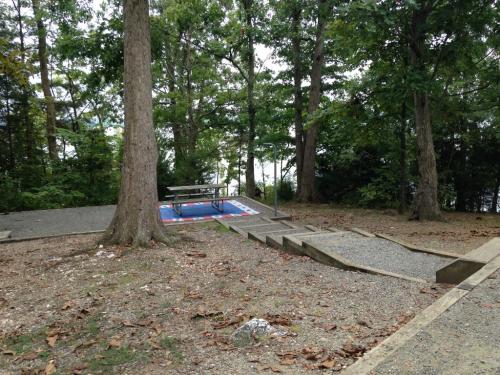 A playground area with a picnic table, a colorful mat, and a gravel path surrounded by trees.
