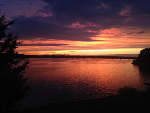A vibrant sunset over a calm river, with colorful clouds reflecting on the water and silhouettes of trees in the foreground.