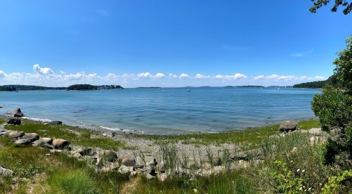 A panoramic view of a calm bay with clear blue water, green grass, and scattered rocks under a bright sky.