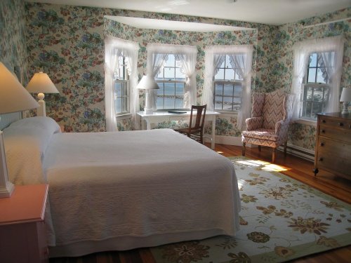 Bright bedroom with floral wallpaper, a white bed, a cozy armchair, and large windows overlooking a scenic view.
