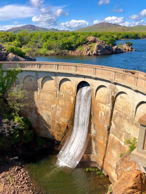 A scenic view of a dam with a waterfall cascading into a serene lake, surrounded by green hills and blue skies.