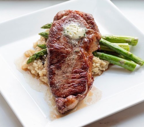 A perfectly cooked steak topped with butter, served with asparagus and creamy risotto on a white plate.