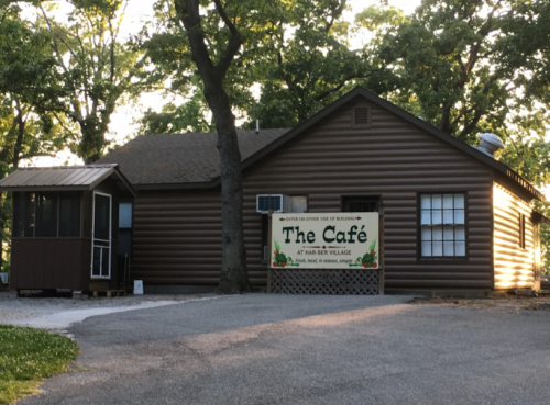 A rustic log cabin with a sign reading "The Café" surrounded by trees and a gravel path.