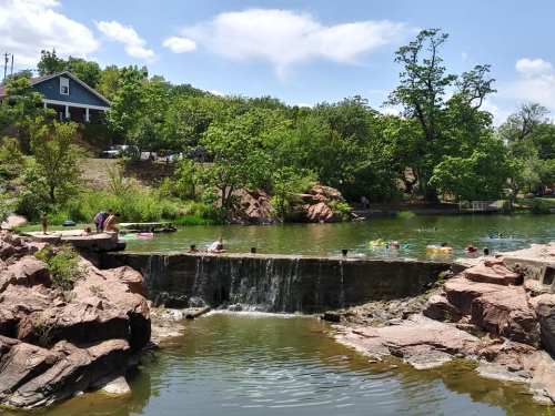 A serene park scene with a small waterfall, people swimming, and lush greenery under a clear blue sky.