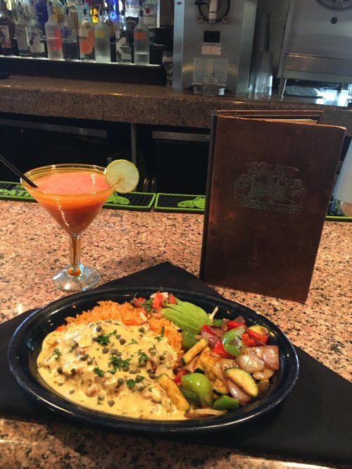 A colorful plate of chicken with cheese sauce, rice, and vegetables, alongside a cocktail and a menu on a bar counter.