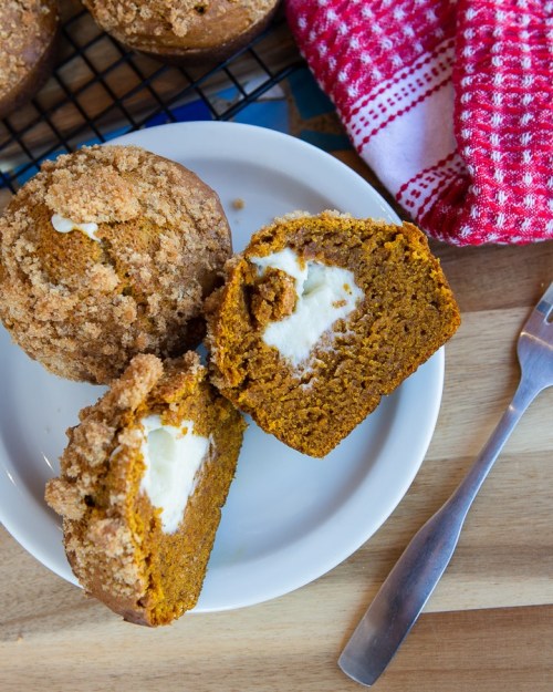 A plate with a pumpkin muffin, cut in half to reveal cream filling, next to a whole muffin and a red checkered napkin.