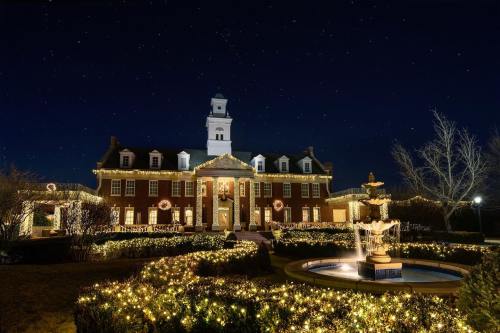 A beautifully lit historic building at night, adorned with holiday decorations and surrounded by twinkling lights and a fountain.