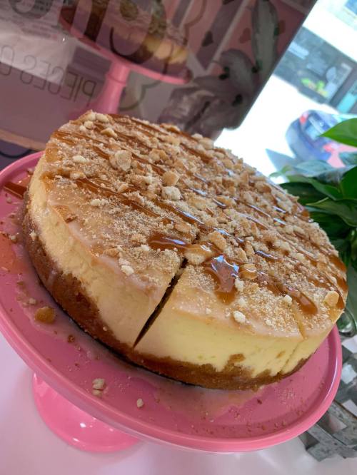 A delicious cheesecake topped with caramel drizzle and crushed cookies, displayed on a pink cake stand.
