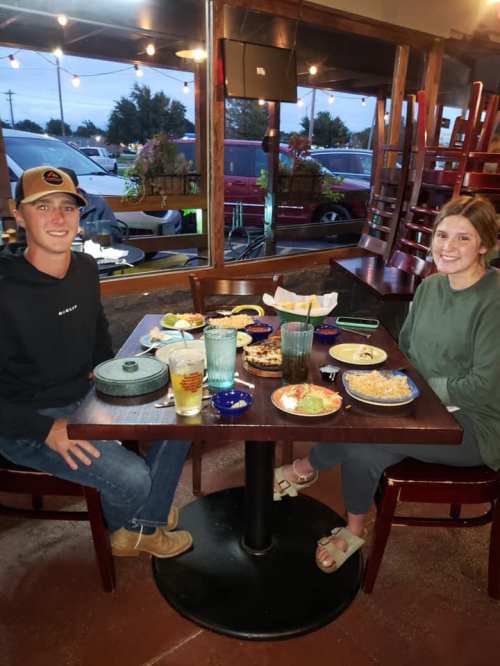 A young man and woman sit at a table filled with food and drinks in a cozy restaurant setting.