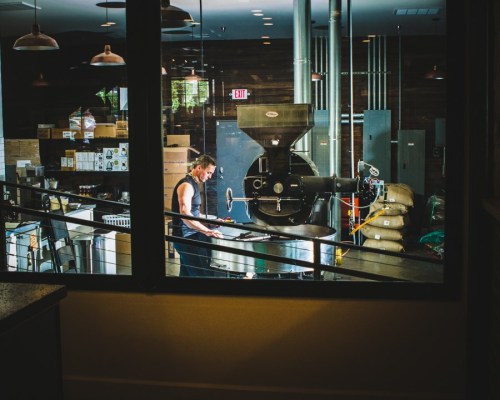 A person working at a coffee roasting machine inside a modern café, with boxes and equipment in the background.