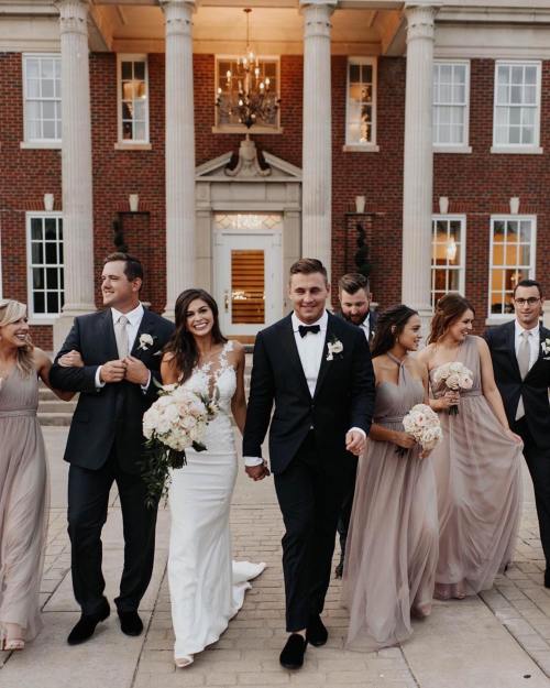 A joyful wedding party walks together outside a grand building, with the bride and groom in the center, holding hands.