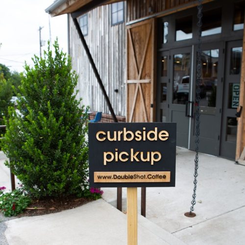 A sign for curbside pickup outside a coffee shop, with a website link and a rustic wooden building in the background.