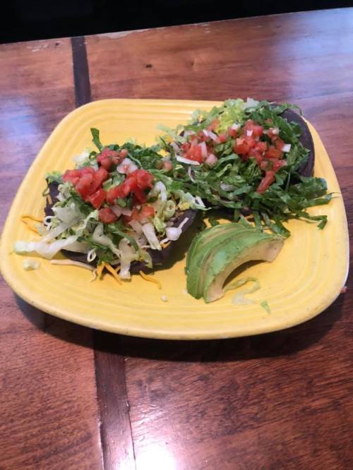 Two colorful tacos topped with lettuce, tomatoes, and cheese, served on a yellow plate with a slice of avocado.