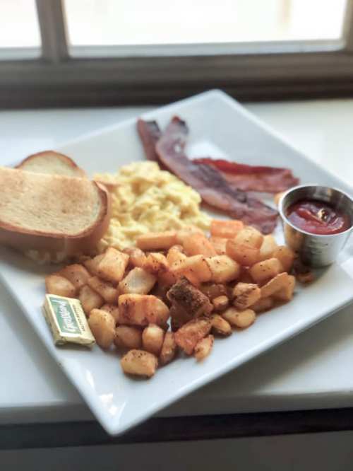 A breakfast plate with scrambled eggs, bacon, toast, seasoned potatoes, and a small cup of ketchup.