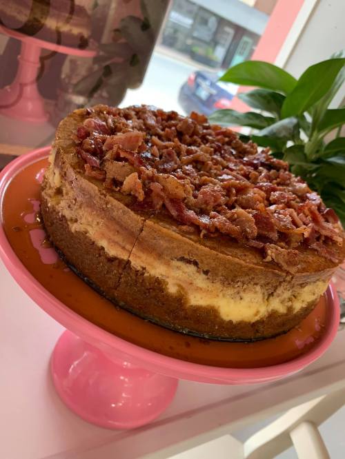 A cheesecake topped with crispy bacon, displayed on a pink cake stand with a plant in the background.