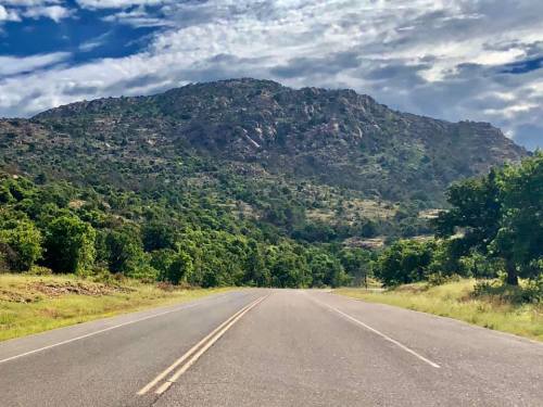 A scenic road leads towards a mountainous landscape under a partly cloudy sky, surrounded by lush greenery.