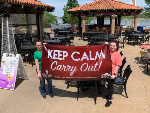 Two people hold a large banner that says "Keep Calm Carry Out!" at an outdoor restaurant by a river.