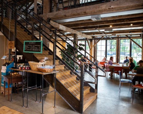 A cozy café interior with wooden beams, a staircase, and patrons sitting at tables, some working on laptops.