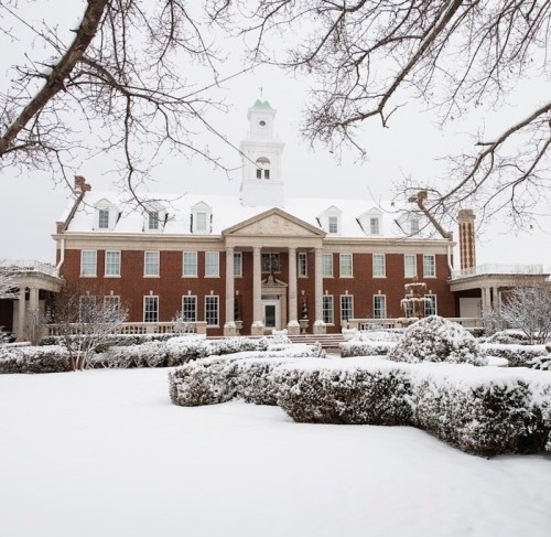 A snow-covered historic building with a clock tower, surrounded by neatly trimmed hedges and bare trees.