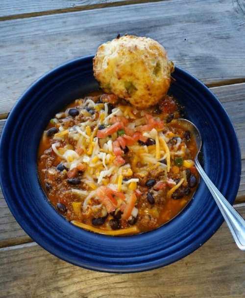 A blue bowl filled with chili topped with cheese and diced tomatoes, alongside a golden biscuit on a wooden table.
