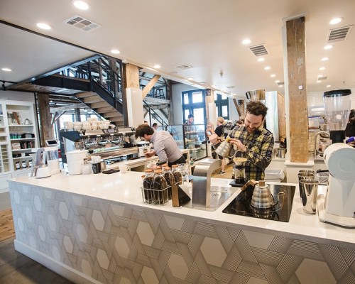 A modern café interior with baristas preparing coffee at a sleek counter and a staircase in the background.