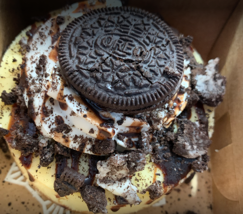 A close-up of a dessert topped with whipped cream, chocolate drizzle, crushed cookies, and an Oreo cookie.