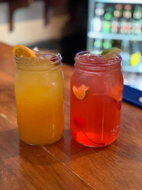 Two mason jars filled with colorful drinks, one orange and the other red, garnished with fruit slices, on a wooden bar.