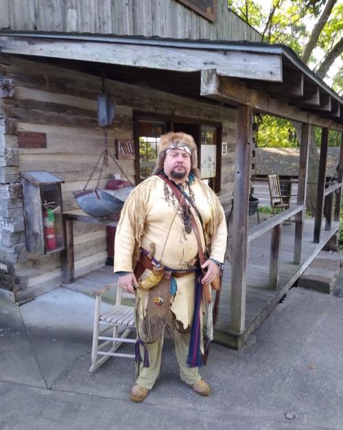 A person in traditional attire stands outside a rustic wooden cabin, surrounded by greenery.