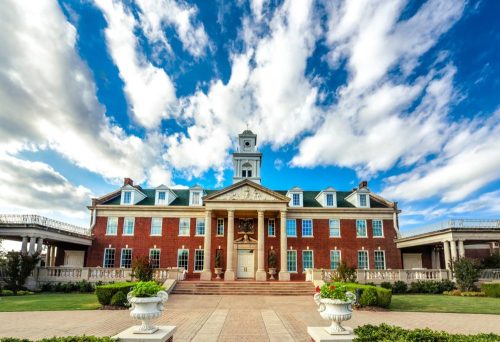 A grand brick building with a clock tower, surrounded by lush greenery and a blue sky with fluffy clouds.