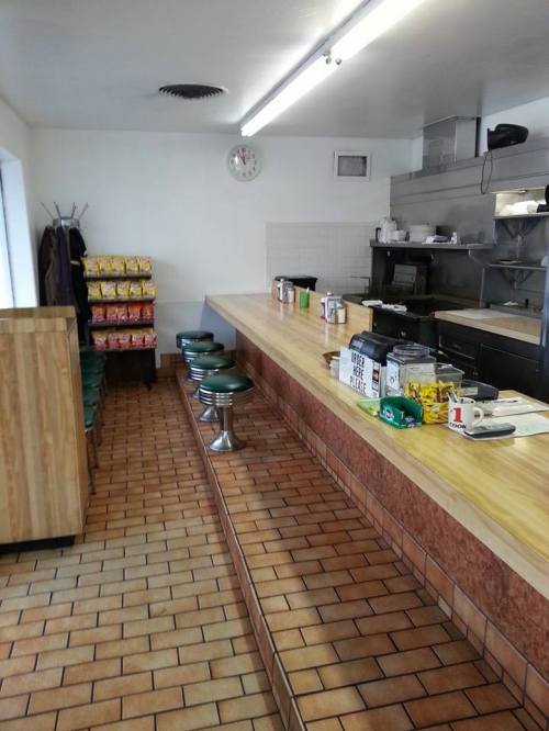 A clean, simple diner interior with a long counter, green stools, and shelves of snacks in the background.