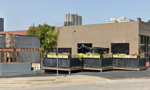 A street view of a building with planters and flowers, surrounded by urban architecture and a clear blue sky.