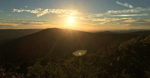 Sunset over rolling mountains, with golden light illuminating the sky and clouds, creating a serene landscape.
