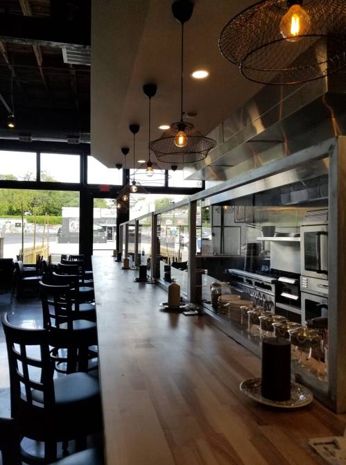 Modern restaurant interior featuring a long wooden counter, pendant lights, and an open kitchen view.
