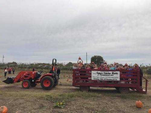 A tractor pulls a wagon filled with people holding pumpkins in a pumpkin patch on a cloudy day.