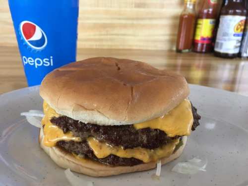 A double cheeseburger with melted cheese and onions, served on a plate next to a Pepsi cup.