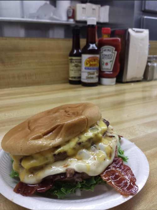 A stacked cheeseburger with bacon, lettuce, and melted cheese on a paper plate, with condiments in the background.