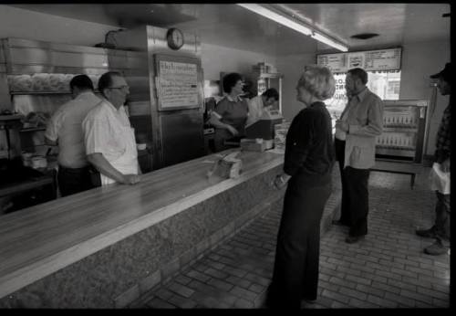 A black-and-white photo of a diner with customers at the counter and staff serving food.