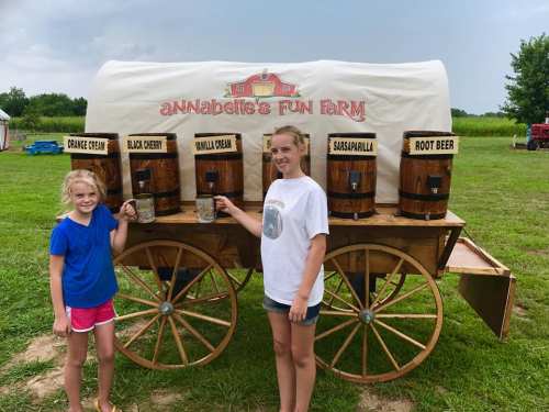 Two girls stand by a wooden cart with barrels labeled for different soda flavors at a farm. Green fields in the background.