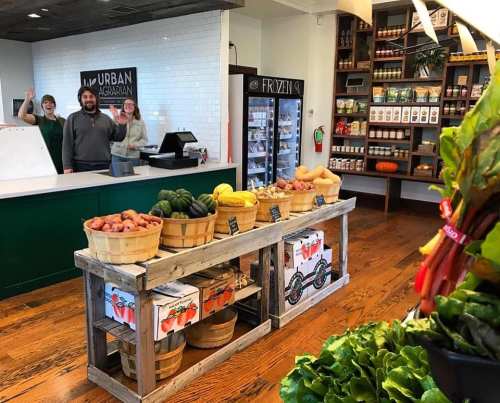 A market scene with staff waving behind a counter, showcasing fresh produce in baskets and shelves of packaged goods.