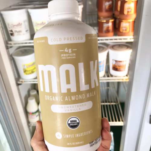 A bottle of MALK organic almond milk in unsweetened vanilla flavor, displayed in a refrigerator with other products.