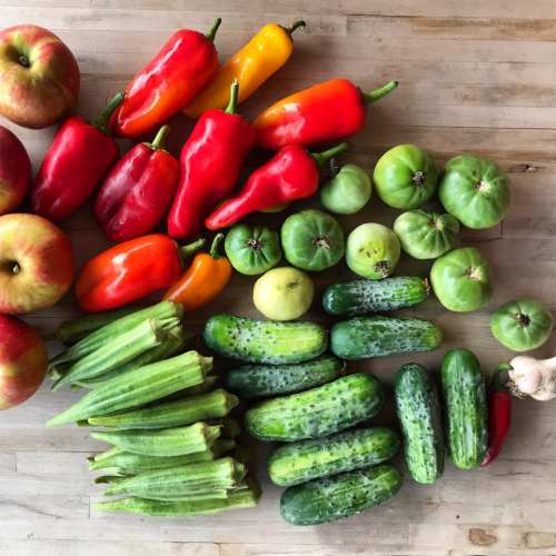 A variety of fresh vegetables and apples arranged on a wooden surface, including peppers, cucumbers, okra, and tomatoes.
