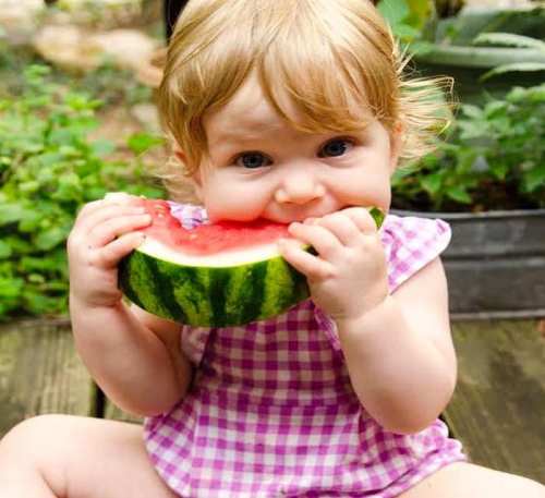 A young child with curly hair sits outdoors, happily biting into a large slice of watermelon.