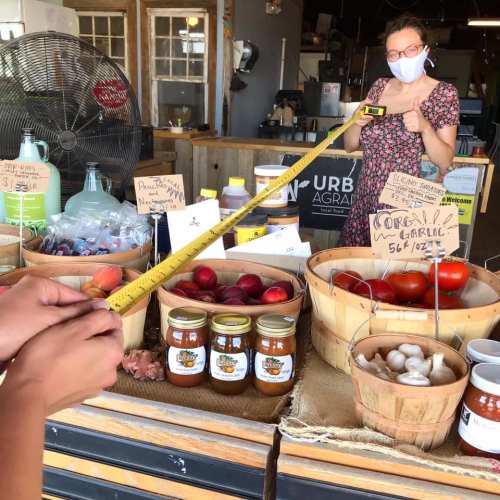A woman in a mask stands behind a market stall with fresh produce and jars, measuring items with a tape measure.