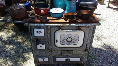 An antique stove with a rustic finish, surrounded by various pots and pans on a gravel surface.