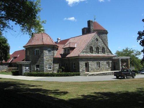 A stone house with a red-tiled roof, featuring a round turret and surrounded by green grass and trees under a blue sky.