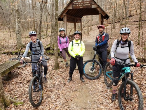 Five mountain bikers pose together on a trail near a wooden shelter in a forested area.