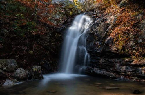 A serene waterfall cascades over rocky cliffs, surrounded by autumn foliage and a calm pool below.